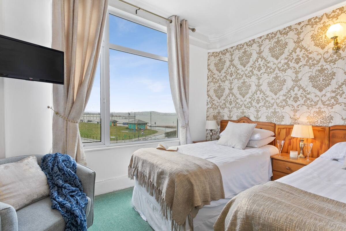 A hotel room with two beds, a TV mounted on the wall, a window with curtains, a patterned wallpaper, and a nightstand with a lamp.