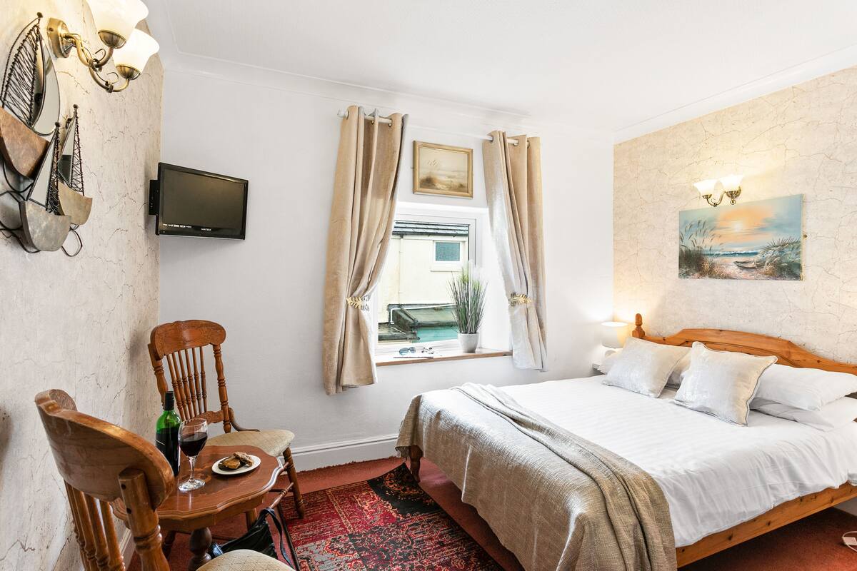 A bedroom with a bed, a chair, a table with a bottle and glass, a TV, and a window with curtains.
