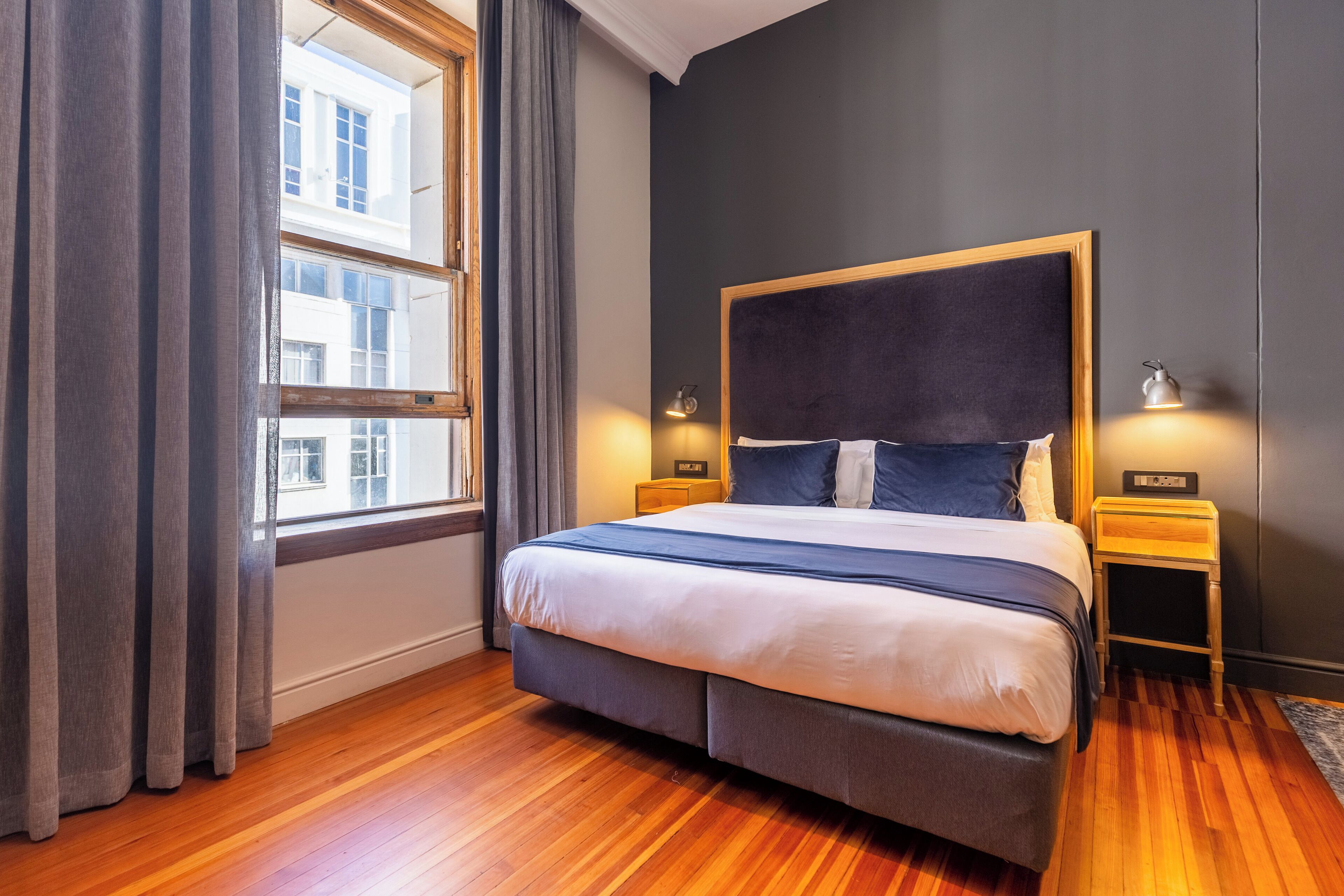 A modern bedroom with a large bed, wooden flooring, and a view of a building through a window.