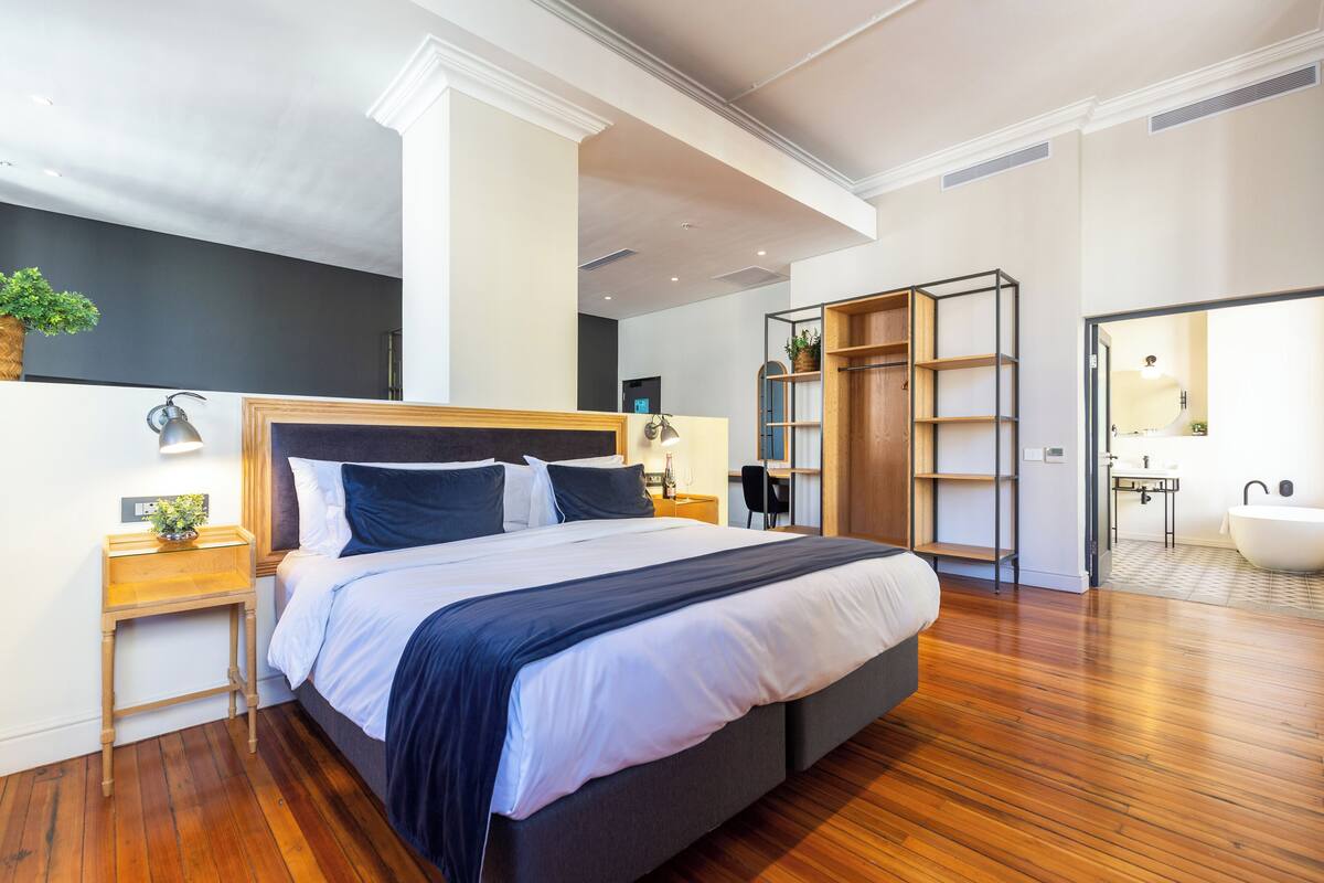 A modern hotel room with a large bed, wooden flooring, a desk, and a bathroom visible through an open door.