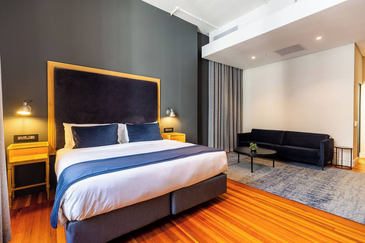 A modern hotel room with a large bed, a sofa, a small table, and a nightstand with lamps.