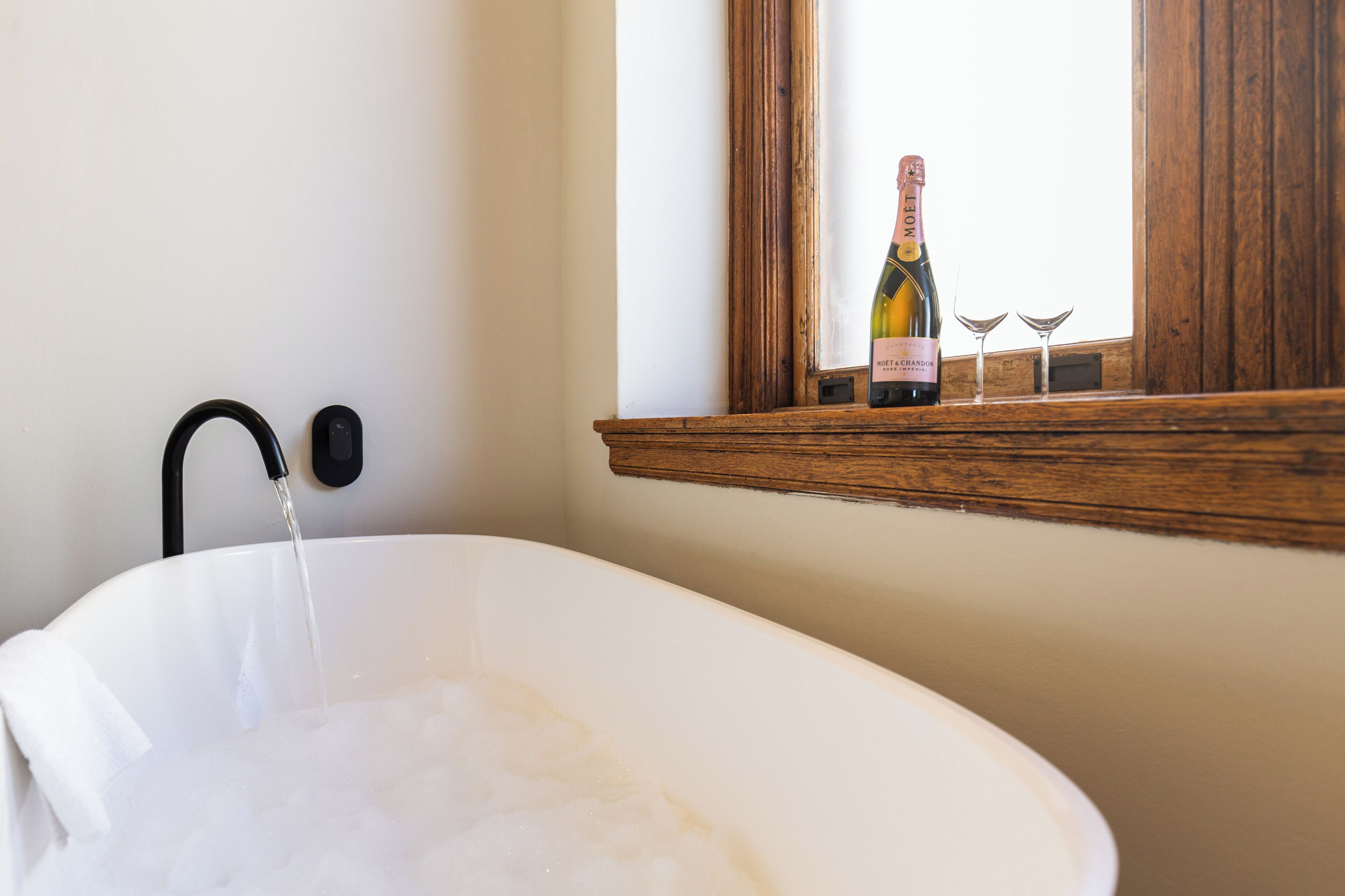 A white bathtub with running water, a bottle of champagne, and two glasses on a wooden shelf by a window.