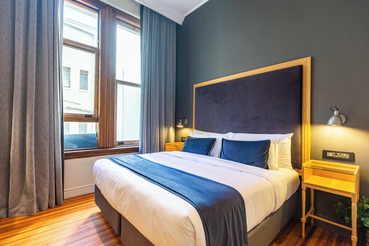 A modern hotel room with a large bed, wooden flooring, a view through a window with curtains, and a bedside table with a lamp.