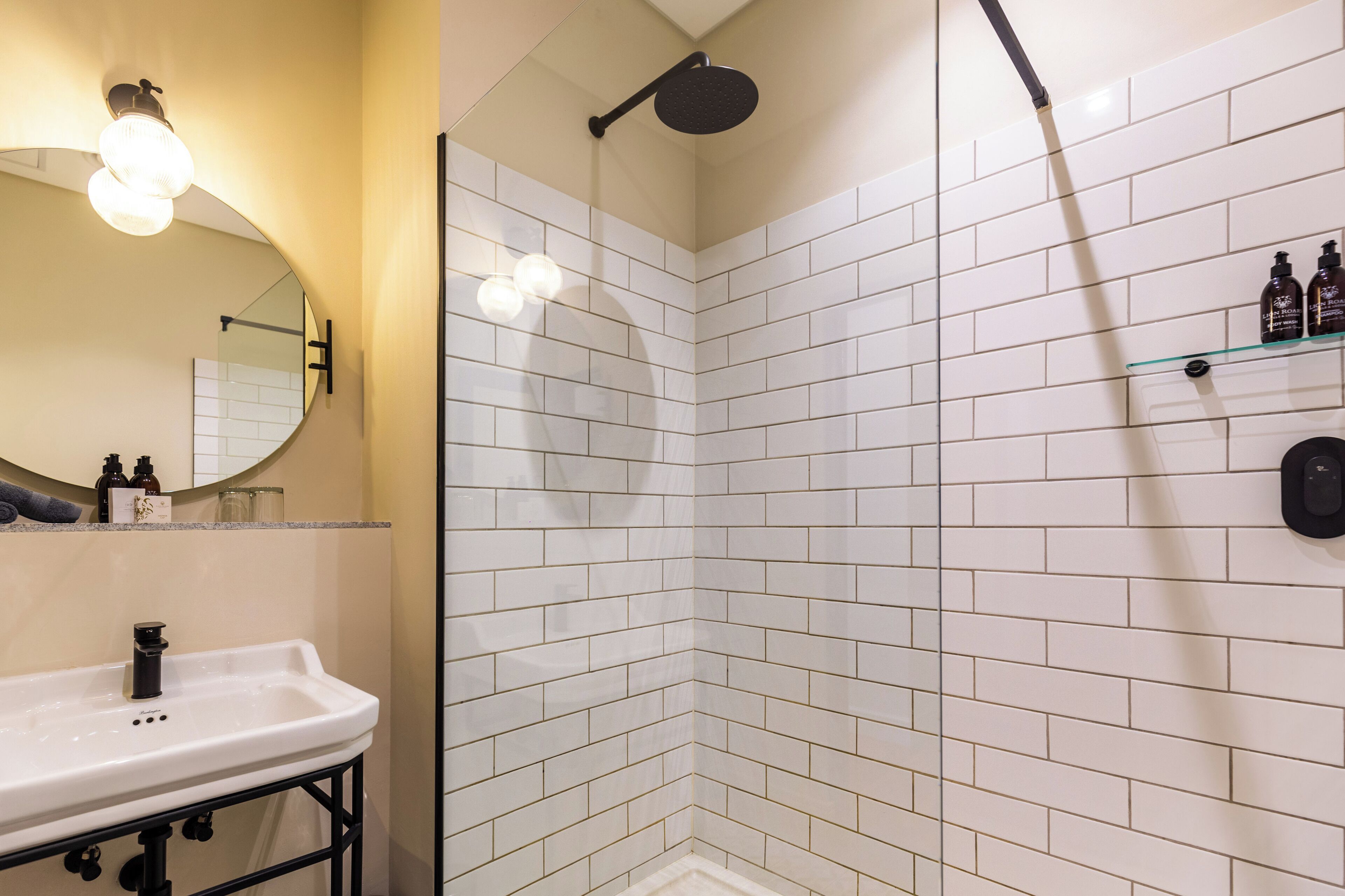 A bathroom with a white-tiled shower, a sink with a black faucet, a mirror, and a shelf with toiletries.