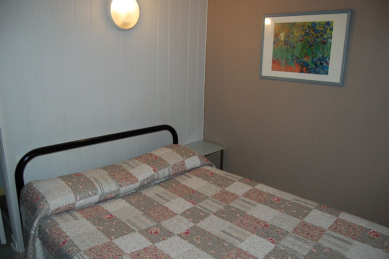 Una cama individual con colcha estampada, una mesita de noche, una lámpara de pared y un cuadro con flores.