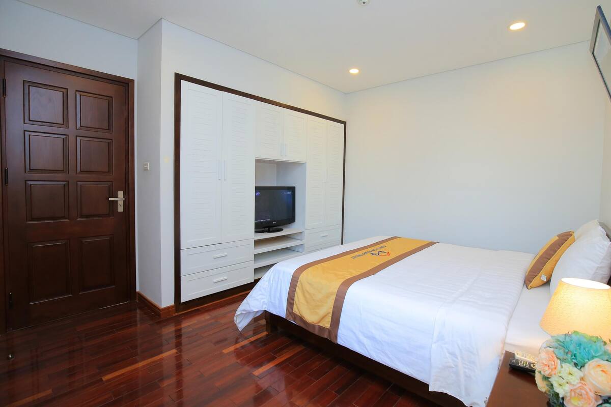 A hotel room with a wooden floor, a bed with white bedding and a yellow accent, a TV in a white entertainment unit, a door, and a wall-mounted light fixture.