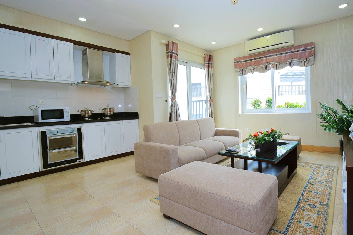A modern living room with a beige sofa, a coffee table, and a TV. There is a kitchen area with a microwave, oven, and cabinets. The room has large windows with curtains and an air conditioning unit on the wall.
