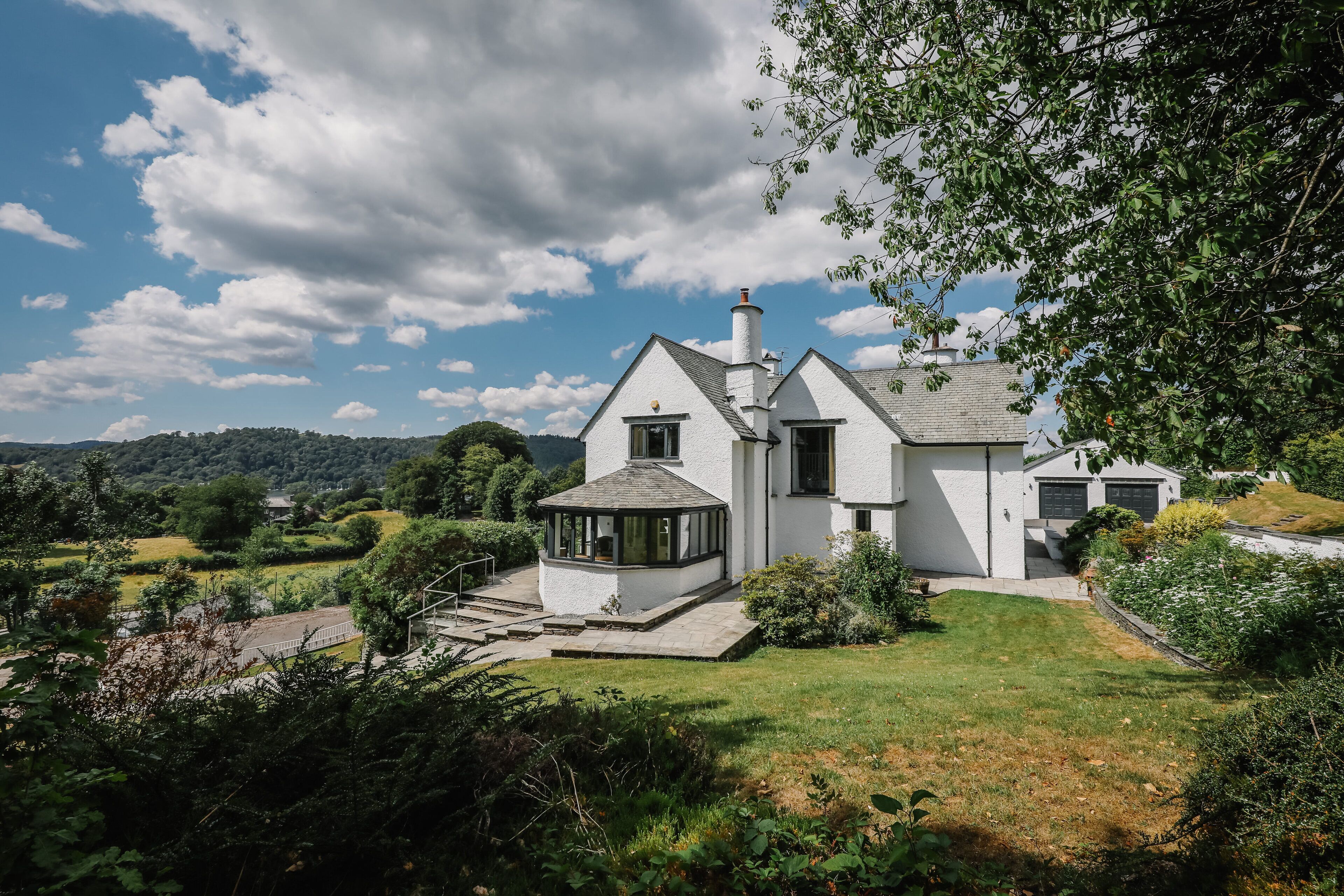 A white house with a large porch and a conservatory, surrounded by lush greenery and hills.