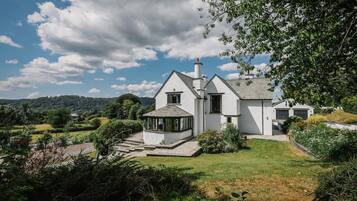 A white house with a large porch and a conservatory, surrounded by lush greenery and hills.