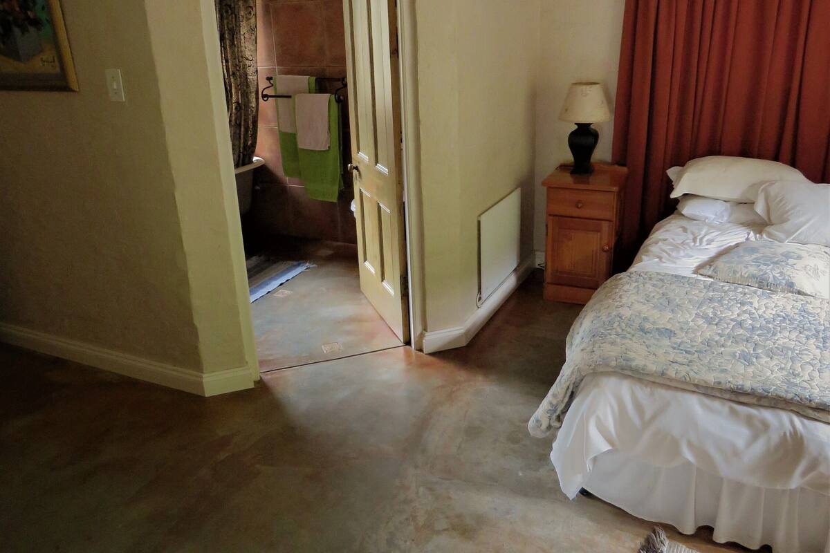 A bedroom with a bed, a nightstand, a lamp, a window with curtains, and a door leading to another room.