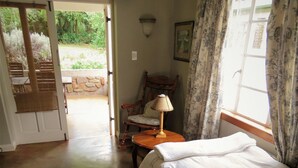 A bedroom with a bed, a chair, a table, and a window with curtains. - Fountain Baths Guest Cottages (Barberton)