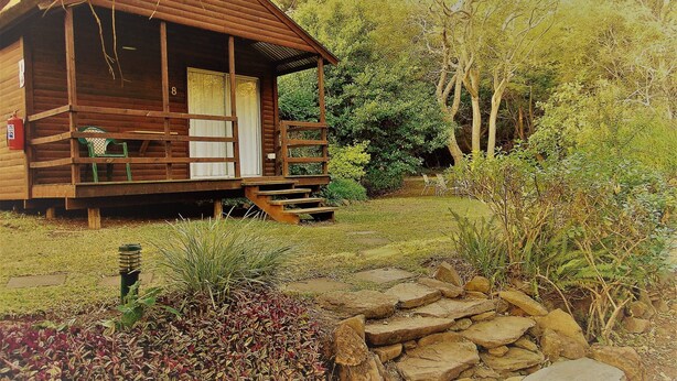A wooden cabin with a porch, surrounded by a garden and trees. - Fountain Baths Guest Cottages (Barberton)