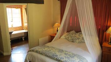 A bedroom with a bed under a canopy, a bedside table with a lamp, a window with curtains, and a small plant on a shelf.