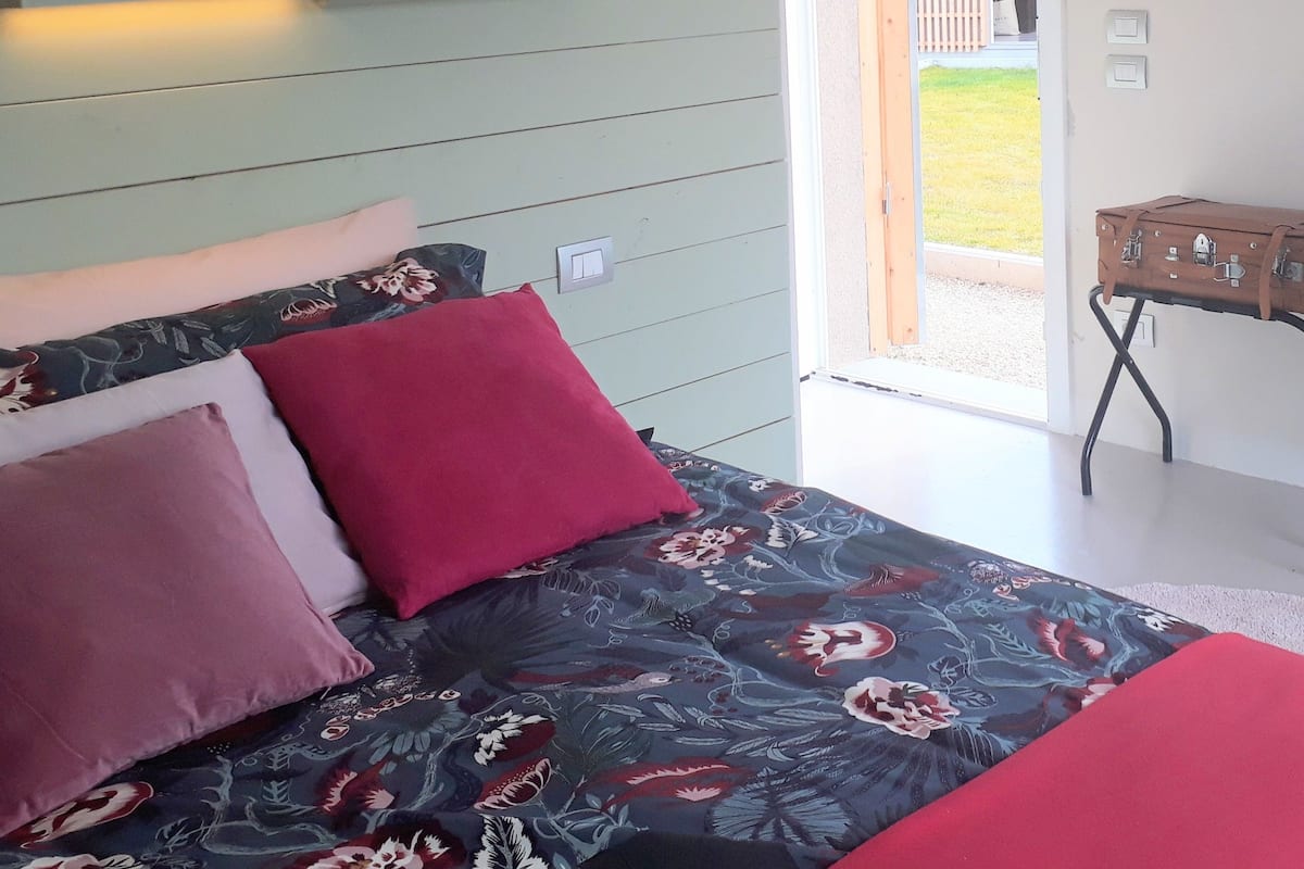 A bedroom with a bed, pillows, a bedside table, a shelf with books and plants, and a view of the outside.