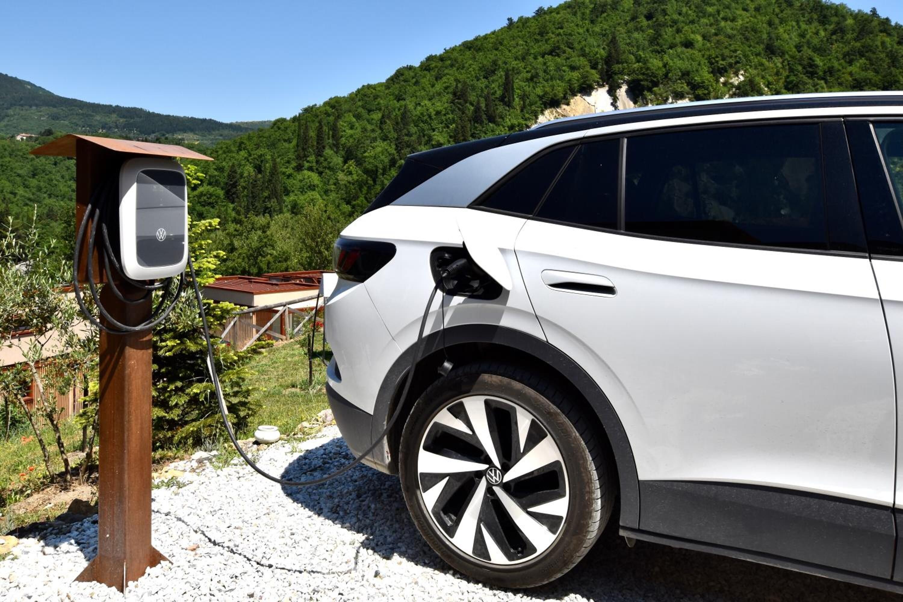 Ein weißer SUV neben einer Ladestation in ländlicher Umgebung, im Hintergrund ein bewaldeter Hügel.