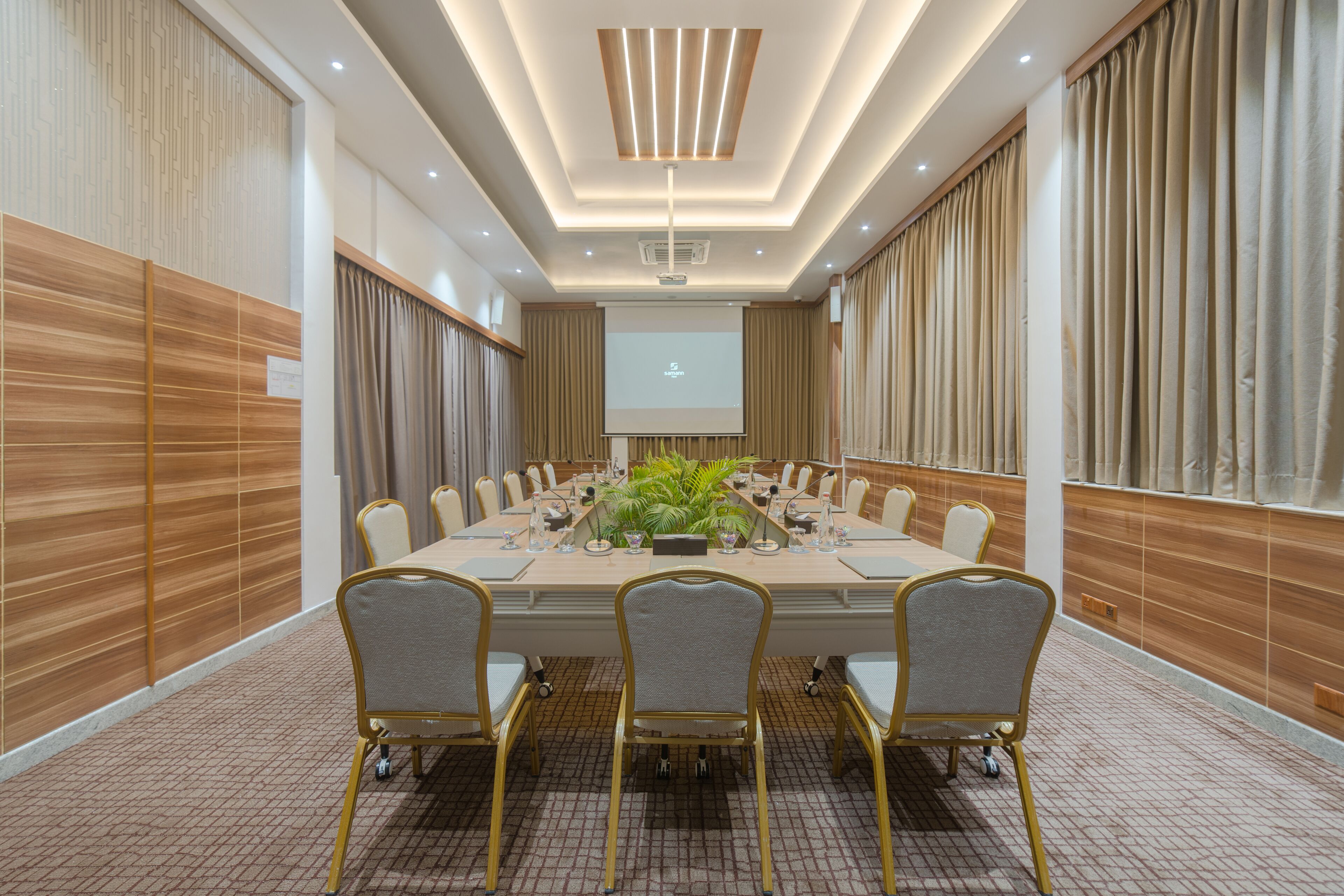 A conference room with a long table, multiple chairs, a projector screen, and a plant centerpiece.