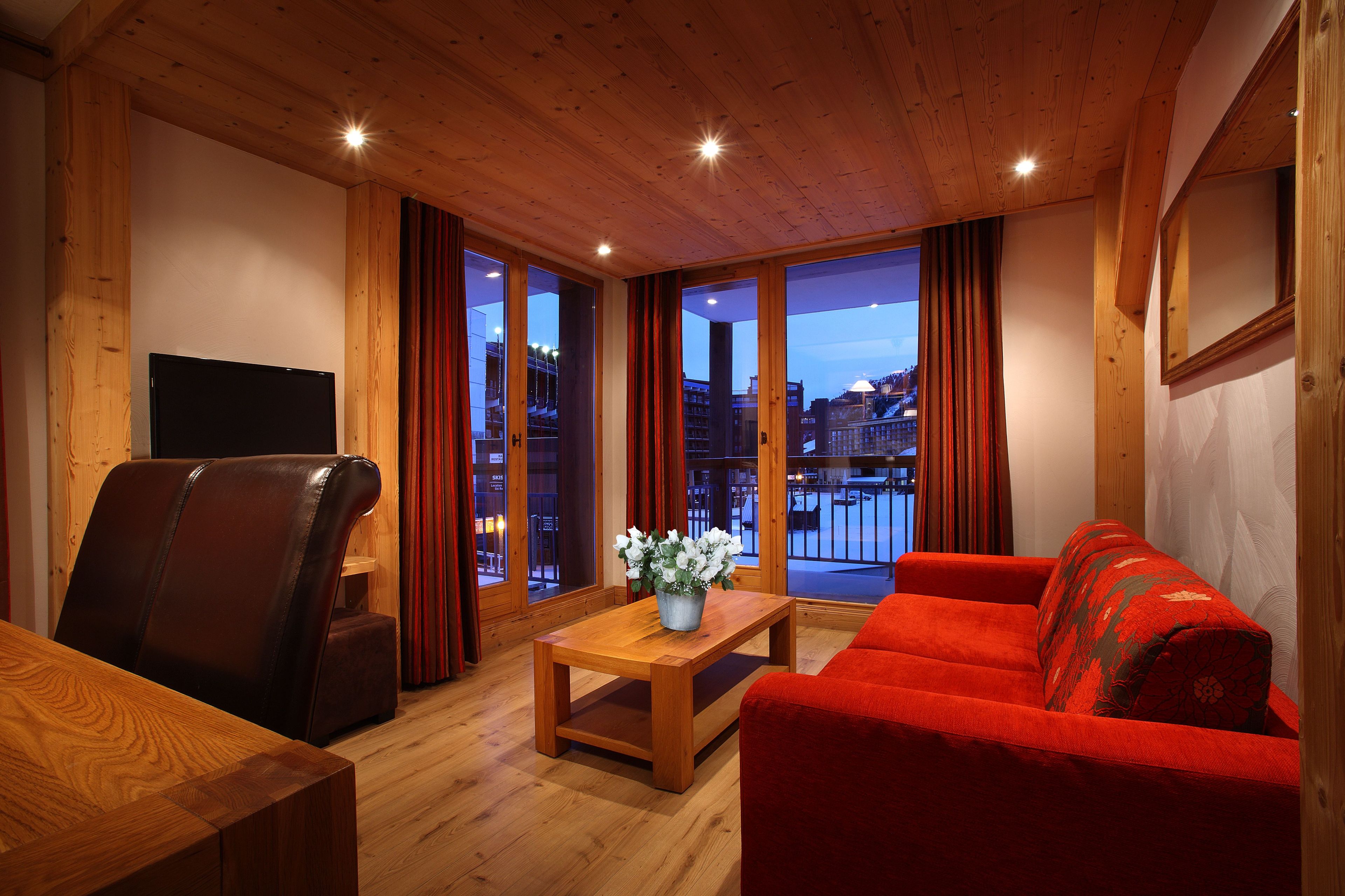 A cozy living room with a wooden ceiling, a leather chair, a red sofa, and a wooden coffee table with a flower vase.