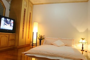 A hotel room with a bed, a TV on a stand, a bedside table with a lamp, and a vase with flowers.