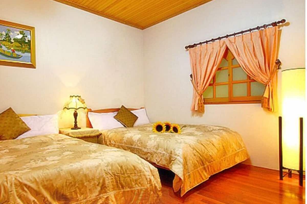A room with two beds, a wooden ceiling, a window with curtains, a lamp, and a painting on the wall.