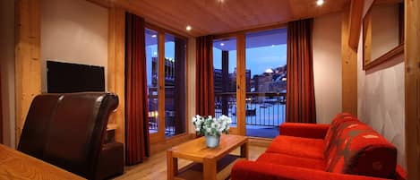 Una habitación acogedora con techo de madera, un sofá rojo, una mesa de centro de madera con un jarrón de flores, un sillón de cuero y un televisor en la pared.