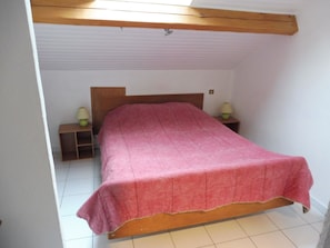 Un dormitorio con cama de madera, colcha rosa, dos mesitas de noche con lámparas y techo inclinado.