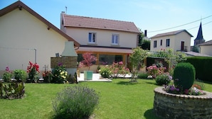 Une maison à deux étages, avec un toit en tuiles, un jardin agrémenté de diverses plantes et fleurs, et un bac à fleurs en pierre.