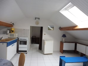 Cocina compacta con microondas, estufa y refrigerador. Hay un pequeño comedor con mesa y sillas. Se observa un reloj en la pared.