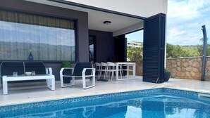 Un área de piscina exterior moderna con una piscina azul cristalina, sillas de descanso blancas y un fondo de pared de piedra.
