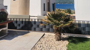 Un edificio moderno con techo plano y balcón. Una palmera está en primer plano, y un cuerpo de agua con montañas al fondo.