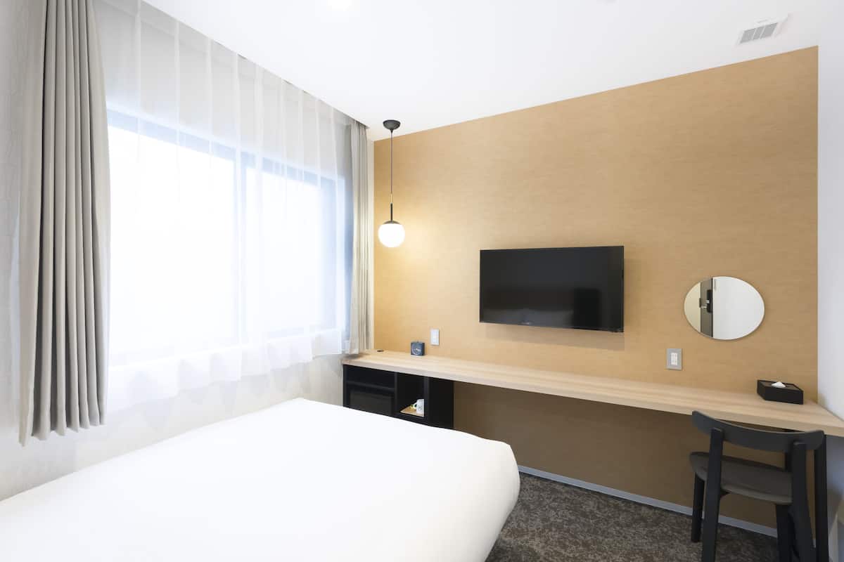 A hotel room with a bed, a desk with a flat-screen TV, a mirror, and a window with curtains.