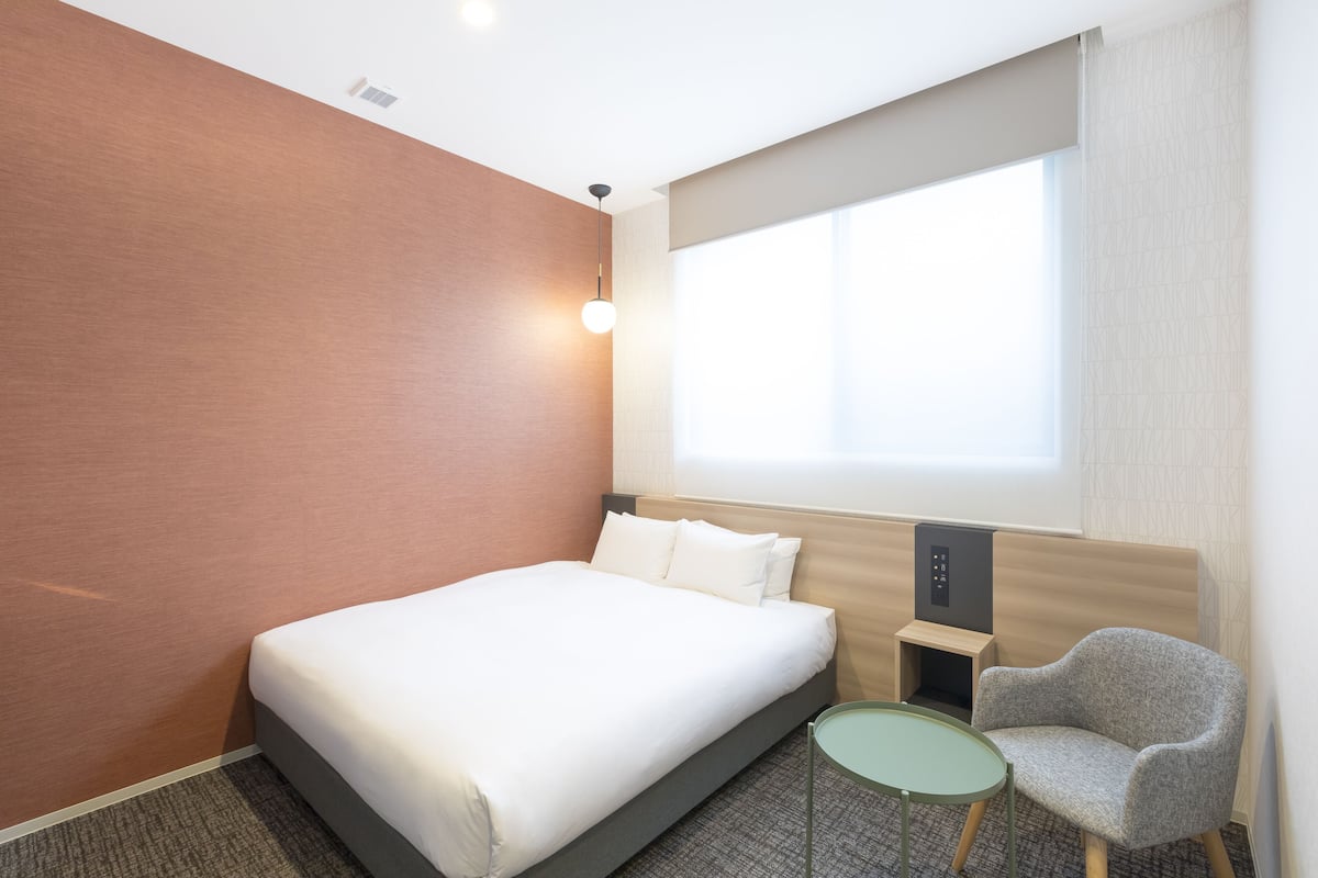 A hotel room with a bed, a small table, a chair, and a window with curtains.