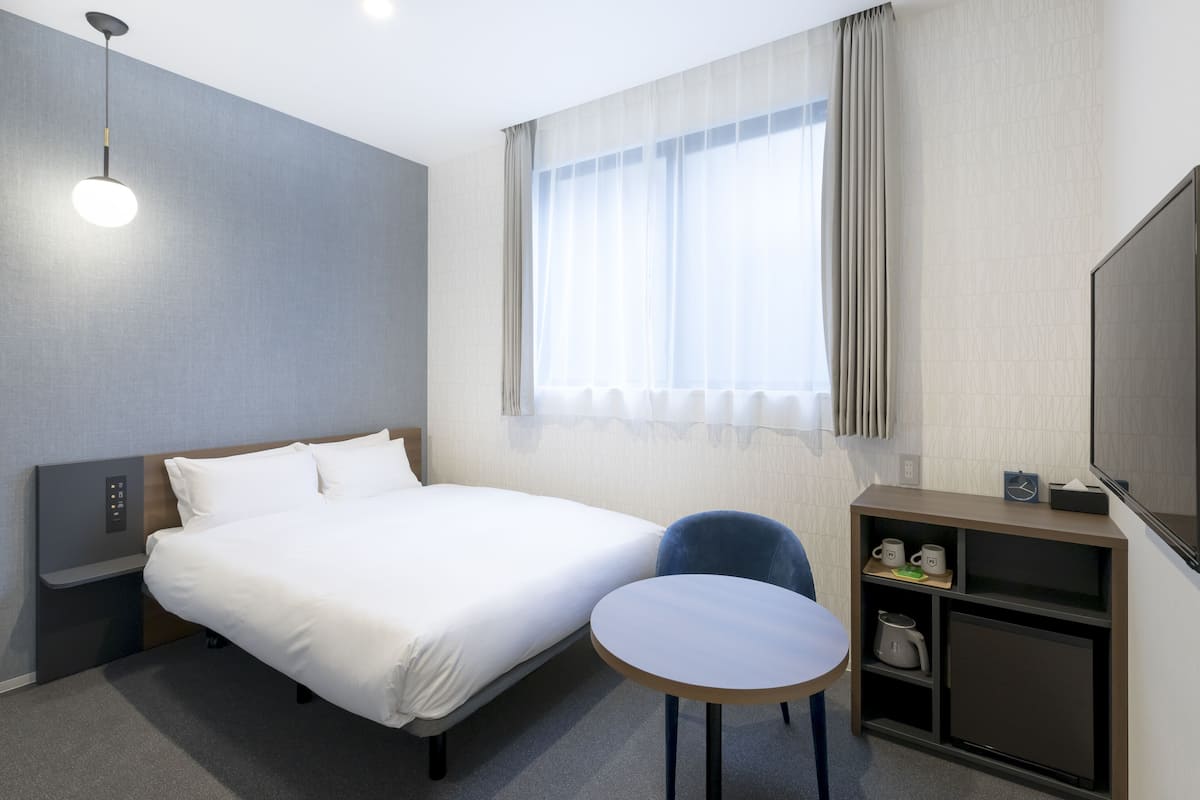 A hotel room with a bed, a small table, a chair, a TV, and a shelf with a kettle and cups.