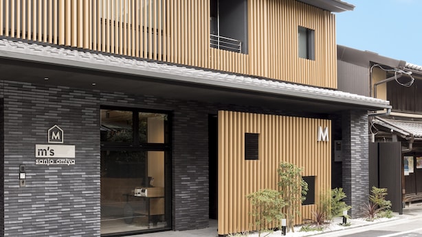 A modern building with a 'M's' sign, featuring a mix of wooden and brick facades, large windows, and a small garden area. - M’s Hotel Sanjo Omiya (Kyoto)