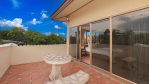 Un balcone con tavolo e panchetta in pietra, una porta scorrevole in vetro che dà su una camera da letto con un letto e una sedia, e un pavimento piastrellato.