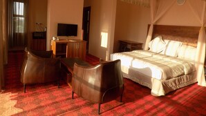 Ein Hotelzimmer mit einem Bett, zwei Ledersesseln, einem Schreibtisch mit Fernseher und einem Fenster mit Vorhängen.