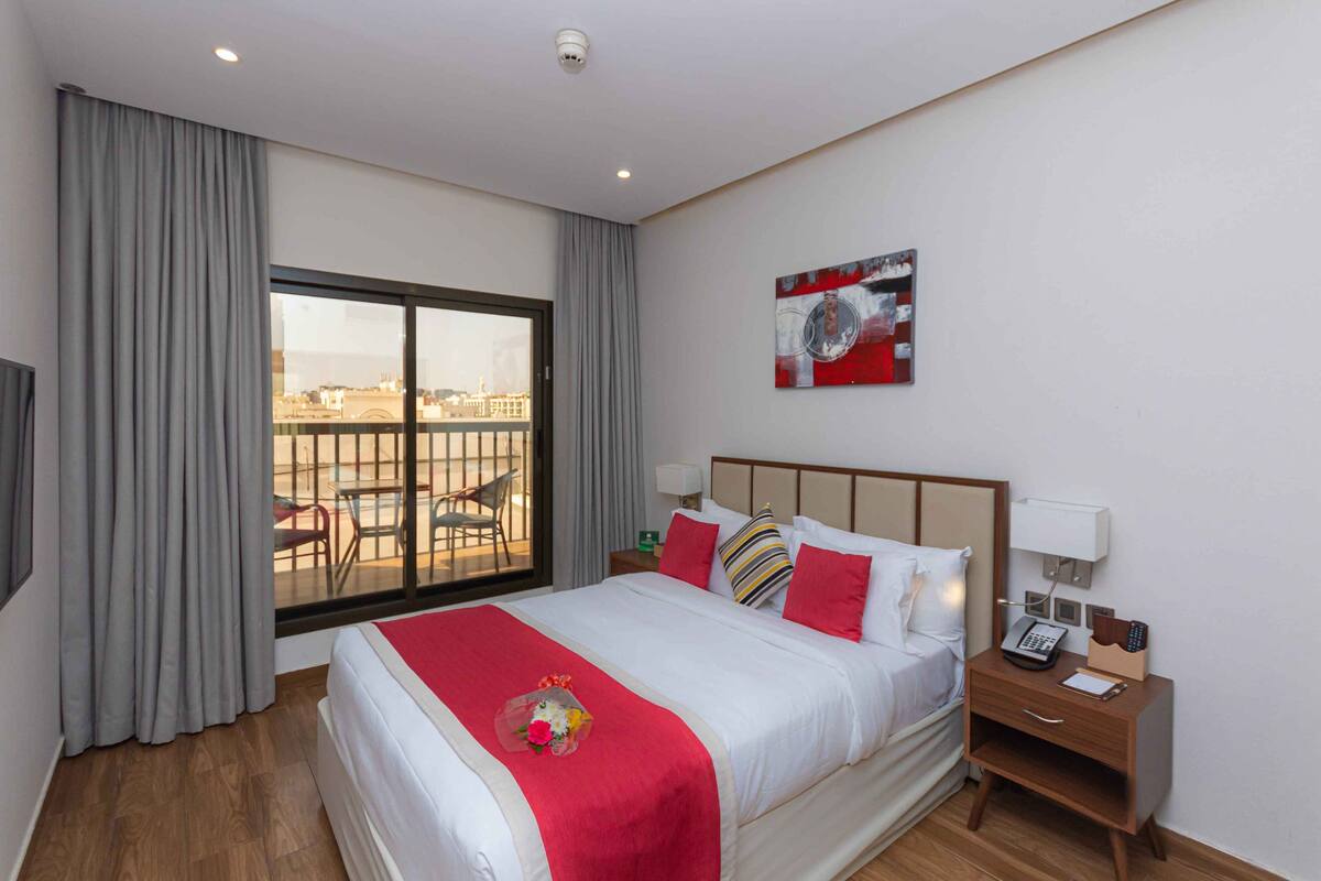 A hotel room with a large bed, a nightstand with a phone and lamp, a view of the city through a sliding door, and a painting on the wall.