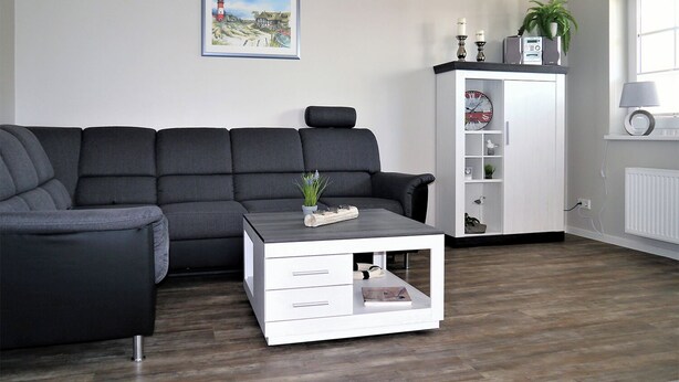 A modern living room with a sectional sofa, a white cabinet with shelves, a small table with decorative items, and a framed picture on the wall. - Centrally located apartment close to the beach for max. 4 pers. with terrace, WLAN, washing machine (Ostseebad Prerow)