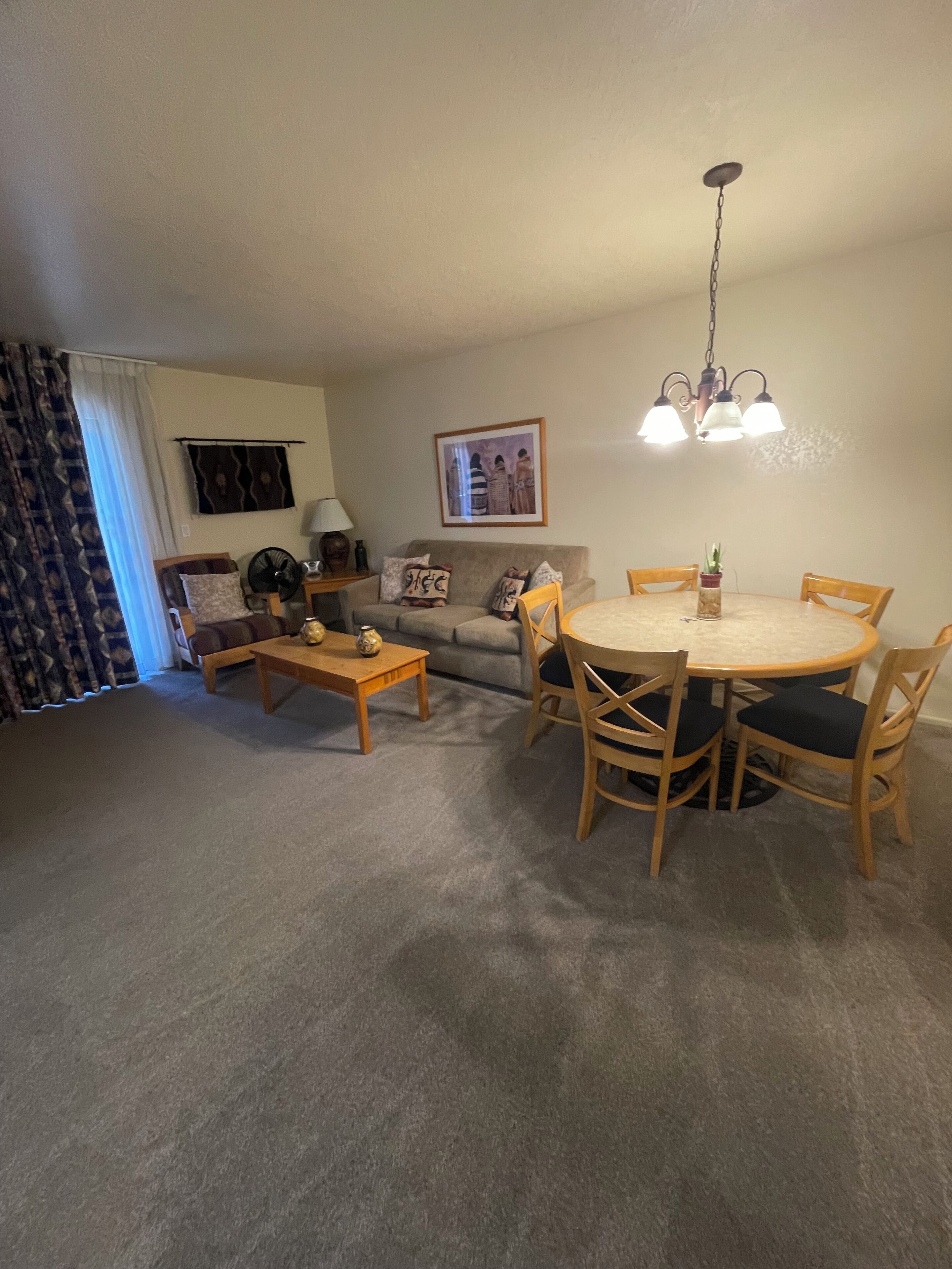 Een woonkamer met een bank, salontafel en een eettafel met stoelen. Er is een plafondventilator, een lamp en een lijst met een afbeelding aan de muur.