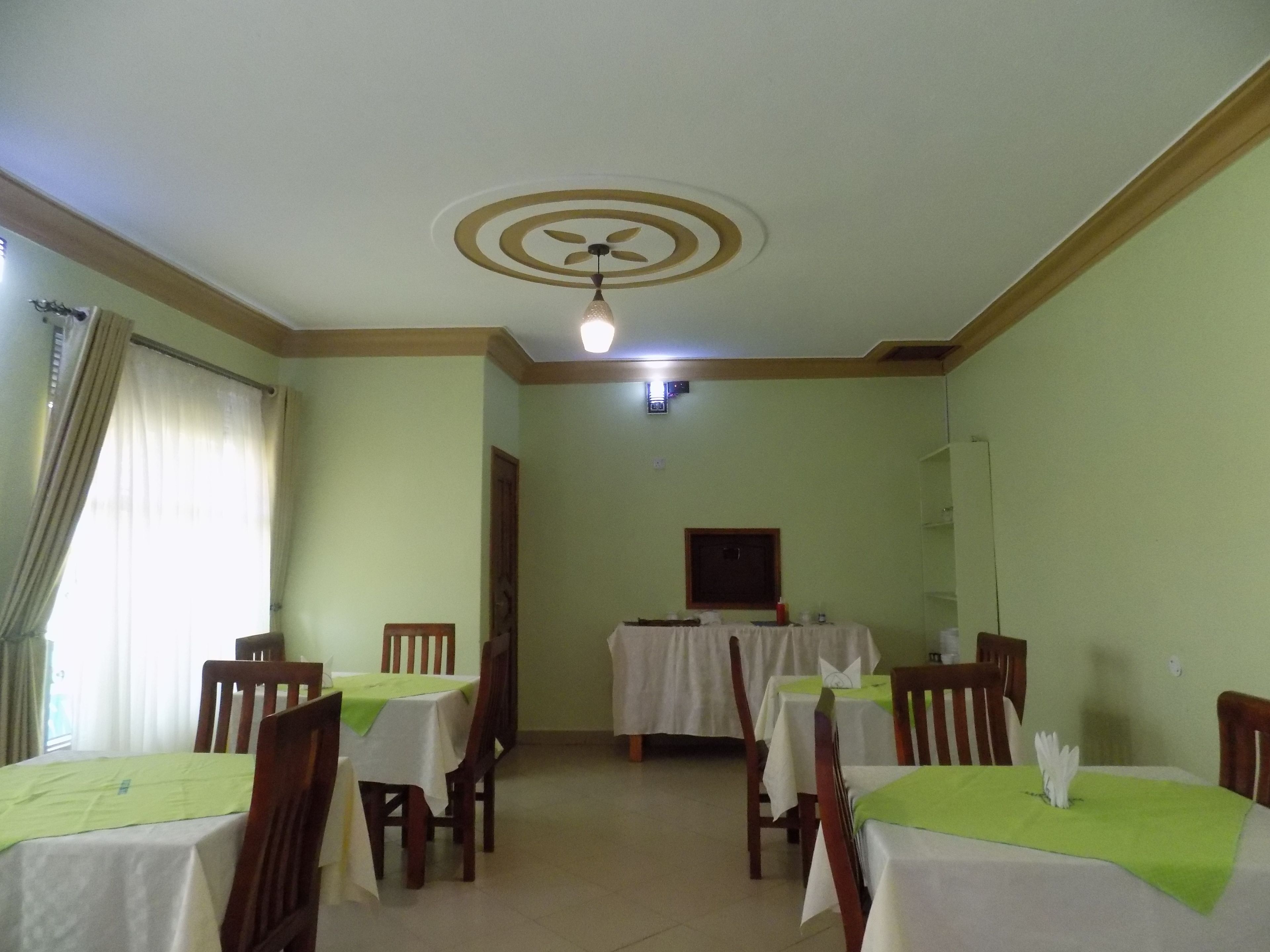 A dining area with tables set for four, each with a green table runner and white tablecloths. There are wooden chairs around the tables. The room has a ceiling fan and a light fixture.