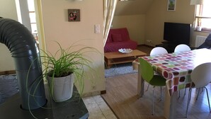 A living room with a sofa, a dining table with chairs, a potted plant, a television, and a window with curtains.