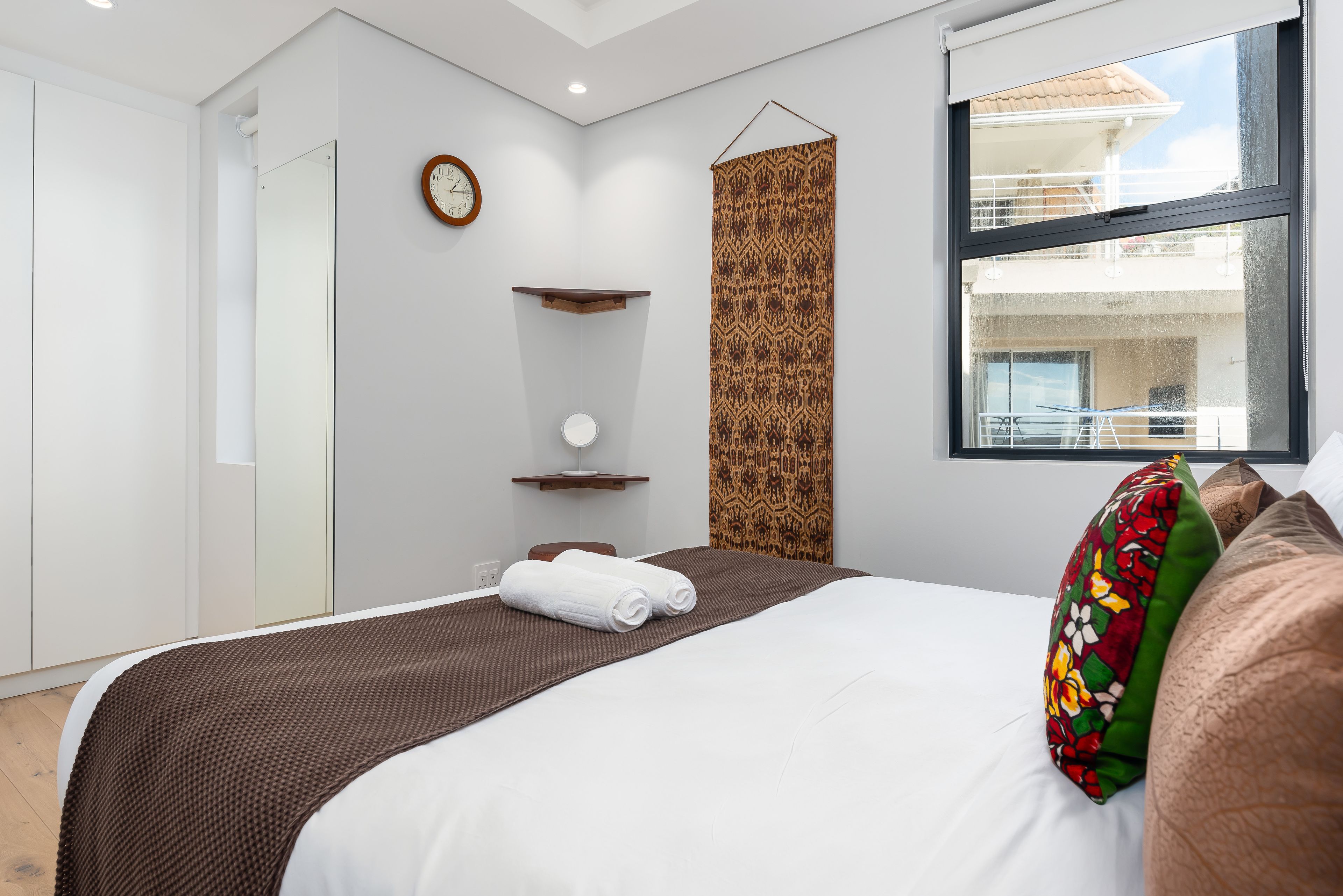 Une chambre d’hôtel moderne avec un grand lit, un rideau décoratif suspendu, une petite étagère avec un miroir rond et une vue sur le balcon extérieur.