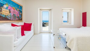 A hotel room with a white bed, a white sofa with red pillows, a painting of a colorful town scene, and a view of the sea through a door. - Villa Coventina (Mykonos)