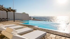 A poolside area with white lounge chairs, a stone floor, and a view of the sea. - Villa Coventina (Mykonos)