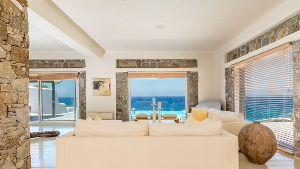 A spacious living room with a large white sofa, stone walls, and a view of the ocean. - Villa Coventina (Mykonos)