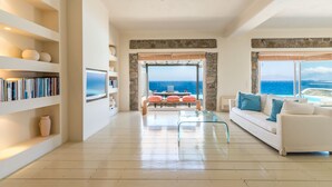 A spacious living room with a large window offering a view of the sea, a white sofa with blue cushions, and a bookshelf filled with books. - Villa Coventina (Mykonos)