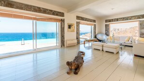 A spacious living room with large windows overlooking a beach, a white sofa, and a large exercise ball. - Villa Coventina (Mykonos)