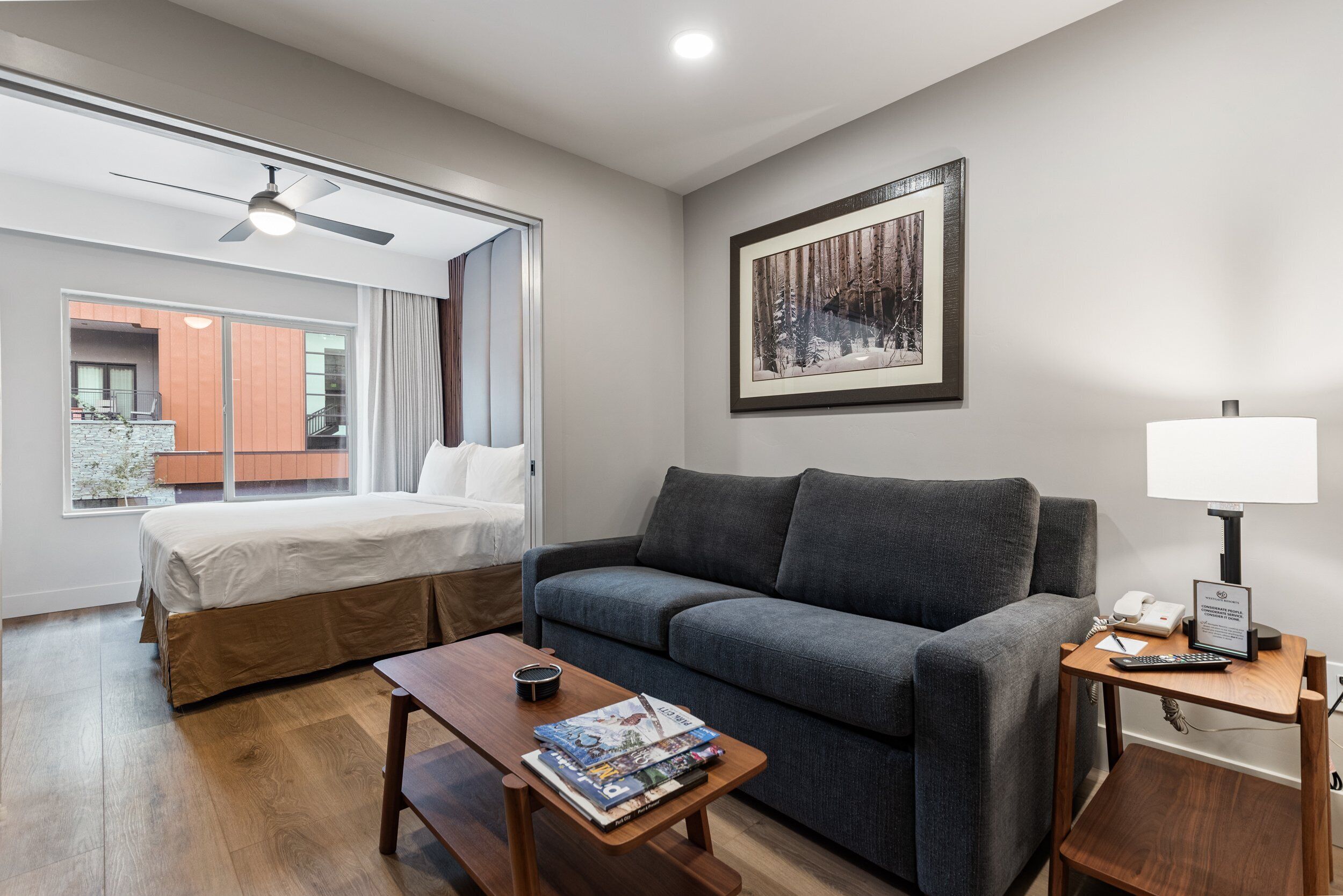 A hotel room with a bed, a gray sofa, a wooden coffee table with magazines, a lamp, and a framed picture on the wall.