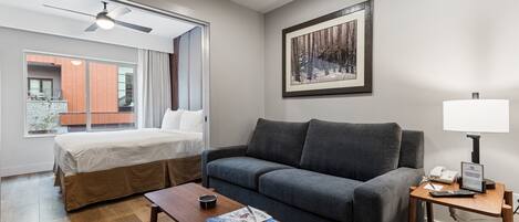 A hotel room with a bed, a gray sofa, a wooden coffee table with magazines, a lamp, and a framed picture on the wall.