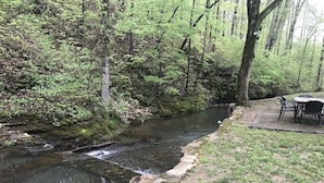 A natural setting with a stream, surrounded by trees and a picnic area with chairs and a table.