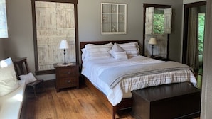 A bedroom with a wooden floor, a bed with white bedding, a nightstand with a lamp, a ceiling fan, and a window with a view of greenery.